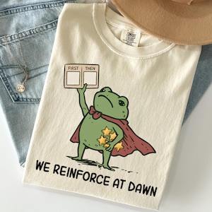 Pode incluir: Uma t-shirt cor de creme com um desenho de um sapo verde com uma capa vermelha, segurando uma placa que diz FIRST THEN e estrelas amarelas. Abaixo do sapo, o texto diz WE REINFORCE AT DAWN. A t-shirt tem uma etiqueta da marca Comfort Colors.
