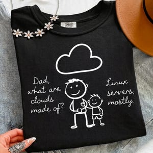 May include: A black t-shirt featuring a white line drawing of a cloud above a parent and child figure. The text reads: Dad, what are clouds made of? Linux servers, mostly. The shirt has a crew neckline and a Comfort Colors brand label inside.