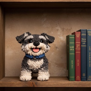 Op de afbeelding: Een handgemaakte gehaakte hondenfiguur met een blauwe halsband, zittend op een houten plank. De hond heeft een grijs-witte vacht, zwarte ogen en een roze tong. Boeken zijn zichtbaar op de plank, waaronder "Moby Dick" en "Pride and Prejudice."
