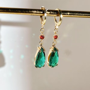 Puede incluir: Un par de pendientes de aro dorados con un diseño colgante. Cada pendiente presenta una pequeña gema roja, una cadena dorada y una gran gema verde en forma de lágrima. Los pendientes se muestran sobre un fondo suave y neutro.