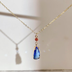 Op de afbeelding: Een goudkleurige ketting met een druppelvormige, blauwe edelsteen hanger. De hanger hangt aan een fijne ketting, met een kleine cluster van rode kralen boven de blauwe steen. De ketting wordt gepresenteerd tegen een zachte, neutrale achtergrond.