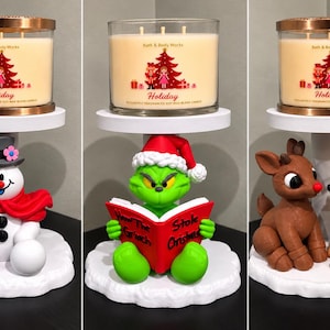 May include: Three Bath & Body Works Holiday candles in glass jars with copper lids, each resting on a white pedestal stand featuring a snowman with a broom, a green character reading a book, or two reindeer figures on a snowy base.