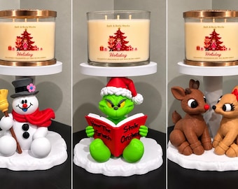 Peut inclure: Trois bougies Holiday de Bath & Body Works dans des bocaux en verre avec couvercles en cuivre, posées sur des socles blancs avec un bonhomme de neige, un personnage vert lisant un livre ou deux rennes sur une base enneigée.