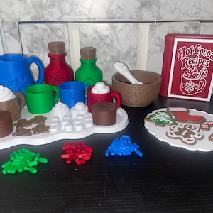 May include: A toy hot cocoa set featuring a blue pitcher, red and green bottles, colourful mugs, a brown bowl with a white spoon, and a red book titled Hot Cocoa Recipes. Includes white marshmallow shapes, brown gingerbread cookies, and small sprinkles.