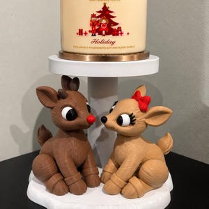 May include: A three-wick Bath & Body Works Holiday candle in a glass jar with a copper lid, featuring a nutcracker and tree graphic. Below, two brown reindeer figurines sit on a white tiered stand, one with a red nose and one with a red bow.
