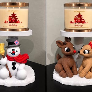 May include: Two Bath & Body Works Holiday candles in glass jars with copper lids, each resting on a white pedestal. One pedestal features a snowman with a broom and red scarf, the other features two brown reindeer with red accents.