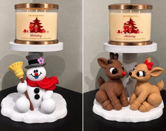 May include: Two Bath & Body Works Holiday candles in glass jars with copper lids, each resting on a white pedestal. One pedestal features a snowman with a broom and red scarf, the other features two brown reindeer with red accents.