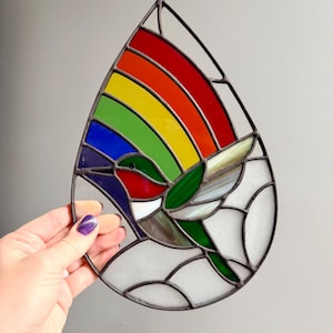 Op de afbeelding: Traanvormige gebrandschilderde glazen suncatcher met een kolibrie en regenboog ontwerp. De regenboog heeft rode, oranje, gele, groene en blauwe segmenten. De kolibrie heeft groene en rode details. Decoratief item.