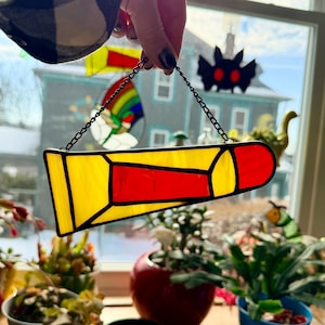 Könnte beinhalten: Ein buntglasförmiger Bleistift-Sonnenfänger mit gelben und roten Paneelen, die schwarz umrandet sind. Er hängt an einer Kette. Der Hintergrund zeigt ein Fenster mit anderen Buntglasarbeiten und Pflanzen.