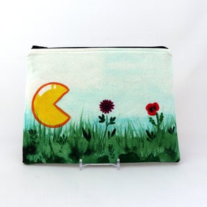 May include: A yellow Pac-Man character with an open mouth is printed on a white canvas zippered pouch. The pouch is surrounded by green grass and two flowers, one red and one purple.