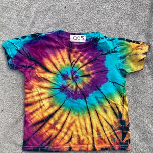 Puede incluir: Una camiseta tie-dye con un patrón en espiral en tonos de morado, azul, amarillo y naranja. La camiseta tiene una etiqueta negra con el número "005" en ella.
