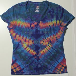 Può includere: Una maglietta con scollo a V tie-dye con un motivo vibrante di tonalità blu, gialle, arancioni e rosse. Il design si irradia dal centro, creando un effetto simmetrico e accattivante. La maglietta è realizzata in tessuto morbido.