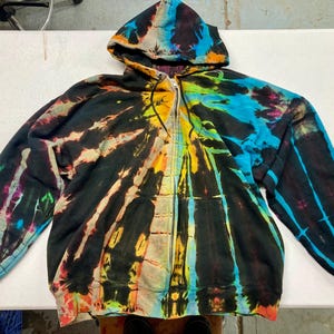 May include: A zip-up hoodie with a vibrant tie-dye pattern. The colors include black, blue, yellow, orange, and purple, creating a spiral design. The hoodie has a hood and two front pockets.