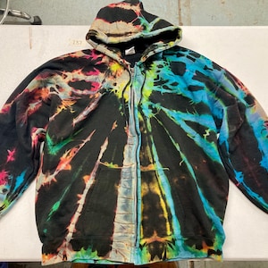 May include: A zip-up hoodie with a vibrant tie-dye pattern. The colors include black, blue, green, yellow, orange, and pink. The hoodie has a hood and a full-length zipper. The sleeves and cuffs are solid colors.