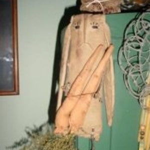 May include: A rustic, handmade bunny-shaped wall hanging made of beige fabric. It features long, floppy ears, a pair of wooden carrots, and a bundle of dried greenery. The bunny hangs from a string, suggesting it's a decorative item.