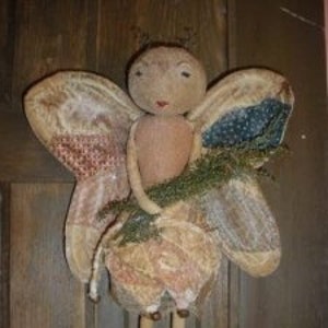 May include: A handmade fabric doll with wings, a floral dress, and a twig in its hands. The doll is made of patchwork fabric in shades of pink, blue, and green. The doll has a simple face with painted eyes and a small mouth.
