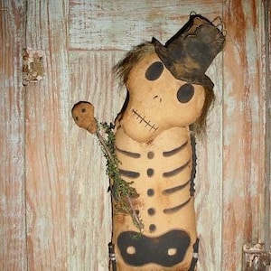 May include: A handmade rag doll skeleton wearing a black top hat and a black eye mask. The doll is made of fabric and has a painted face and skeleton bones.