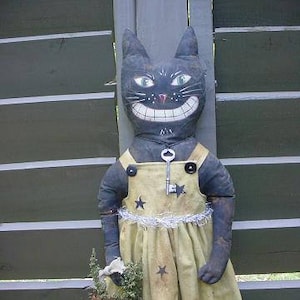 Peut inclure: Une poupée de chat noir avec un sourire narquois porte une robe jaune avec des étoiles noires et un charme de clé argentée. La poupée est debout devant une clôture en bois.