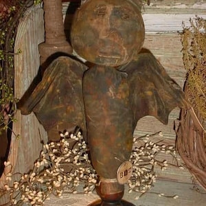 Op de afbeelding: Een rustiek, handgemaakt, bruin en groen vleermuisbeeldje met een geschilderd gezicht en vleugels. De vleermuis staat op een houten voet met een klein label waarop "GHOST" staat.