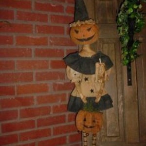 Peut inclure: Une décoration d'Halloween faite à la main représentant une poupée de chiffon avec une tête de citrouille, portant une robe noire et un chapeau pointu. La poupée tient une petite citrouille et une bougie.