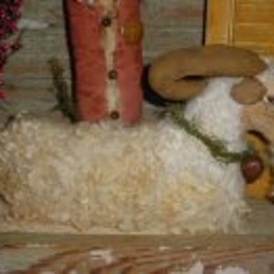 May include: A handmade felt goat, white with brown accents, wearing a green garland with a red heart charm. The goat is sitting on a wooden surface.