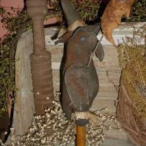 Może przedstawiać: Lalka czarownicy w stylu prymitywnym z czarnym ciałem z tkaniny, brązowym filcowym kapeluszem i drewnianą podstawą. Lalka jest ozdobiona rdzawo-metalicznym wykończeniem.