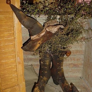 Może przedstawiać: Czarny but czarownicy z ostrym noskiem i czarny kapelusz czarownicy z ostrym wierzchołkiem. But i kapelusz są wykonane z tkaniny i mają rustykalny, vintage wygląd. But jest ozdobiony kokardą z juty, a kapelusz jest ozdobiony wstążką z juty. But i kapelusz stoją na drewnianej półce z kratkowaną tkaniną na pierwszym planie.