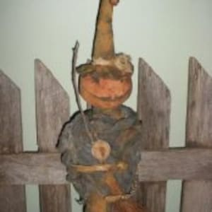 Puede incluir: Una muñeca de bruja primitiva hecha a mano con cabeza de calabaza, sombrero marrón y escoba. La muñeca lleva un vestido marrón y azul y tiene una pequeña calabaza naranja en la mano.