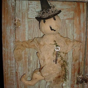 Pode incluir: Um fantasma de pano feito à mão com um chapéu de bruxa preto e uma chave pendurada no seu corpo de estopa. O fantasma é apresentado numa porta de madeira com um acabamento rústico.