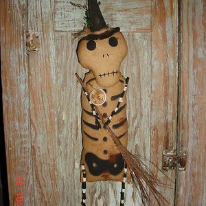May include: A handmade rag doll skeleton wearing a black witch hat and holding a broom. The doll is made of fabric and has a painted face with black eyes and a stitched mouth. The doll is standing on a wooden surface.