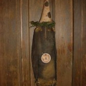 May include: A handmade primitive rag doll with a cone-shaped hat with white polka dots. The doll is dressed in a brown fabric with a white circle with the letter "R" in the center. The doll is hanging on a wooden wall.