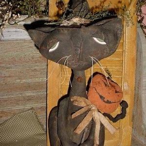 May include: A black cat with a witch's hat and a pumpkin in its hand. The cat is made of fabric and has a rustic, primitive style.