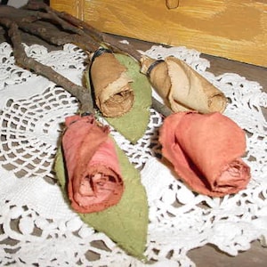 May include: Handmade fabric roses in peach, tan, and pink, attached to brown branches with green fabric leaves. The roses rest on a white lace doily, creating a rustic, vintage aesthetic. A yellow wooden surface is in the background.