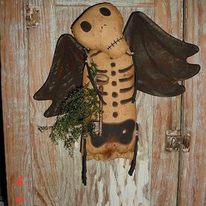 May include: A handmade rag doll with a skeleton design, brown wings, and a key in its hand. The doll is hanging on a wooden door.