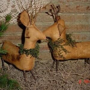 Peut inclure: Deux figurines de cerfs primitives faites &agrave; la main avec des corps en tissu brun et des bois en brindilles. Les cerfs portent des couronnes en tissu vert autour du cou et se tiennent sur un lit de mousse s&egrave;che brune.