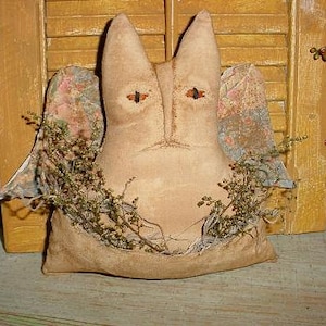 May include: A handmade primitive style fabric doll with a cat-like face and wings. The doll is made of beige fabric and has a floral print on the wings. The doll is sitting on a wooden surface with dried greenery around it.