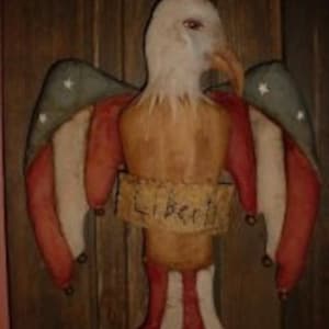 Può includere: Un'aquila patriottica con ali rosso, bianco e blu e uno stendardo che recita "Libertà". L'aquila è realizzata in tessuto e ha uno stile rustico e primitivo.