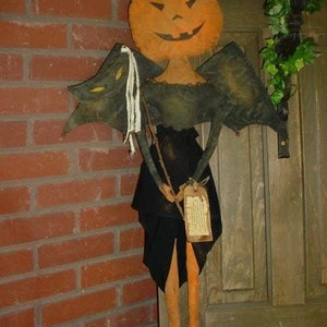 Op de afbeelding: Een handgemaakte Halloween-decoratie met een pompoenkoppop met zwarte vleugels en een zwarte jurk. De pop houdt een stok vast en heeft een label aan zijn taille.