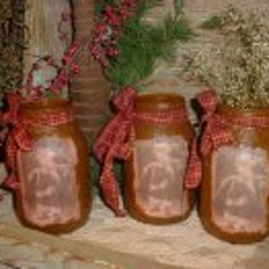 Op de afbeelding: Drie bruine glazen potten met rode en witte geruite linten. Elke pot heeft een wit silhouet van een kerstboom erin. De potten zijn van binnenuit verlicht, waardoor een warme gloed ontstaat.