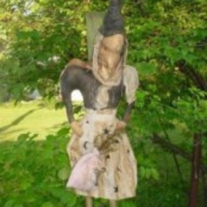 May include: A whimsical witch figurine with a pointed hat, a long brown dress, and a pink bag. The figurine is made of fabric and has a rustic, handmade look.