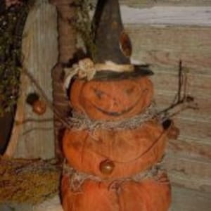 Puede incluir: Una rústica decoración de Halloween con tres calabazas naranjas apiladas que forman una figura similar a un muñeco de nieve. La calabaza superior lleva un sombrero de bruja negro. Ramas y una cadena se utilizan para los brazos y un elemento decorativo.