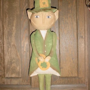 Puede incluir: Un muñeco de duende verde y marrón con un sombrero verde y un trébol en la mano. El muñeco está de pie frente a una puerta de madera.