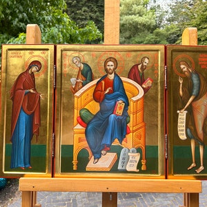 May include: A triptych icon depicting Jesus Christ seated on a throne, flanked by two figures. The central panel shows Jesus holding a book, while the figures on either side appear to be holding scrolls. The icon is painted in a traditional style with gold leaf accents.