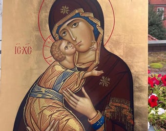 May include: A religious icon featuring a depiction of the Virgin Mary holding the Christ Child. The artwork is rendered in warm tones of gold, brown, and blue, with Greek lettering and a red halo. The icon is displayed on an easel outdoors.