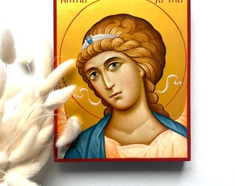 May include: A religious icon featuring a serene angel with light brown curly hair, a blue robe, and a halo. The icon has text in a foreign language at the top and is framed in red. The background is gold.
