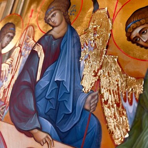 Könnte beinhalten: Ein religiöses Ikonenbild, das drei Figuren mit Heiligenscheinen darstellt. Die zentrale Figur trägt eine blaue Robe und hat einen goldenen Heiligenschein. Die Figuren sitzen an einem Tisch mit einer Schale darauf. Der Hintergrund ist gold und rot.
