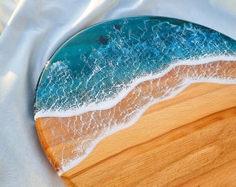May include: A round wooden serving board featuring a decorative resin inlay that mimics an ocean scene. The design includes a light brown wood grain section transitioning into a deep blue and turquoise resin area with white foam waves.