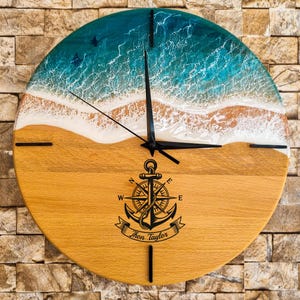 Könnte beinhalten: Runde Wanduhr aus Holz mit Strandmotiv. Die obere Hälfte zeigt einen türkisfarbenen und weißen Ozean, die untere Hälfte einen Sandstrand. Eine schwarze Anker- und Kompassgrafik ist auf dem Zifferblatt zentriert.