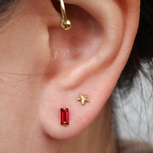 May include: A person's ear with three gold earrings. The top earring is a hoop with a ball at the end. The middle earring is a small gold star. The bottom earring is a red rectangular stone.