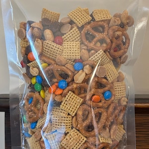 Puede incluir: Una bolsa de plástico transparente llena de una mezcla de aperitivos. La mezcla contiene pretzels, cuadrados de cereal, cacahuetes y caramelos de colores.